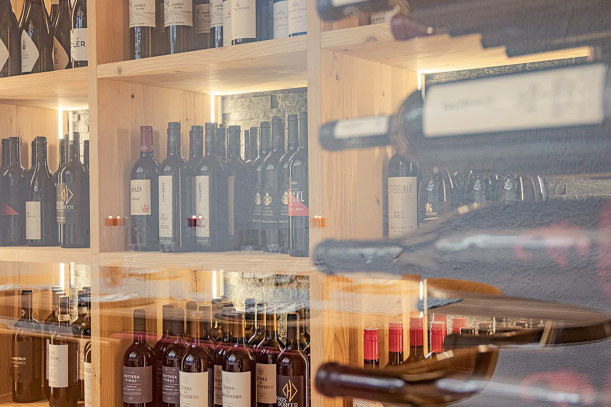 A view of wine bottles on a wooden shelf, partially reflected through a glass partition