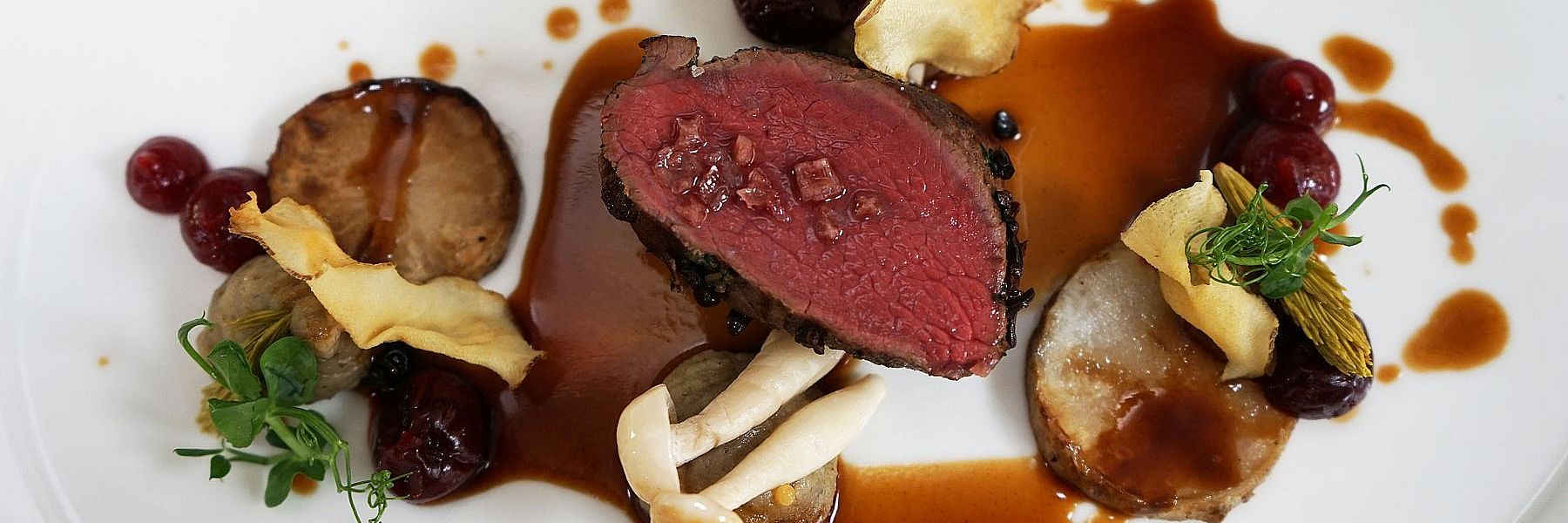 Zartes Wildgericht mit Jus, Wurzelgemüse, Beeren und Pilzen kunstvoll auf weißem Teller angerichtet