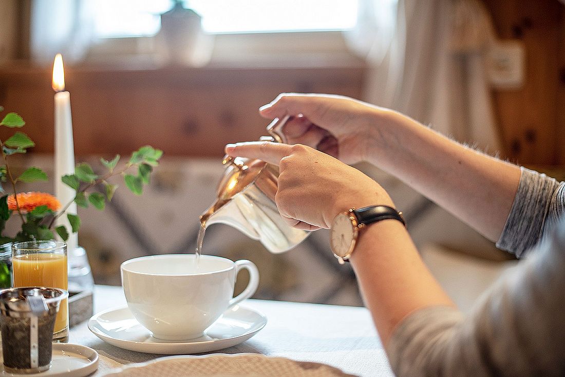 Frau gießt Tee aus silberner Kanne in eine Tasse auf einem gedeckten Frühstückstisch