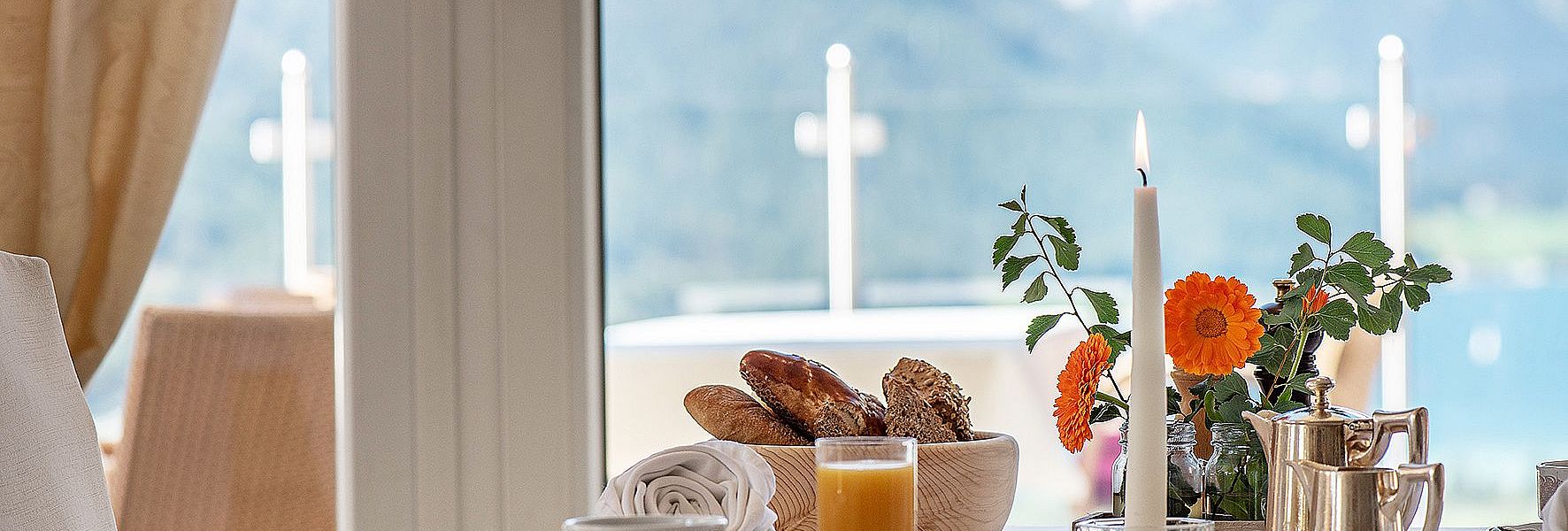 Frühstückstisch mit Gebäck, Saft und Kerze vor Panoramafenster mit Bergblick